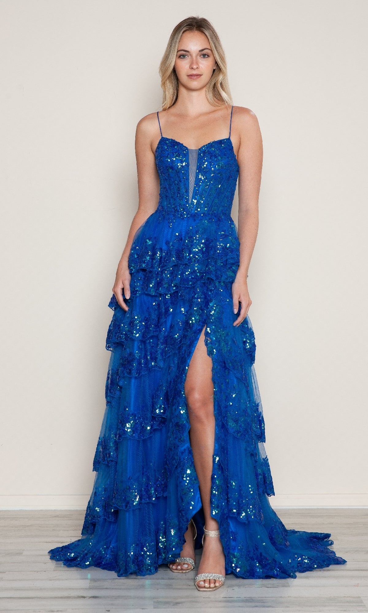 A woman models the Long Prom Dress: Poly USA 9606, featuring blue sequins, a tiered A-line silhouette, thigh-high slit, and spaghetti straps, paired with open-toe heels against a plain background.